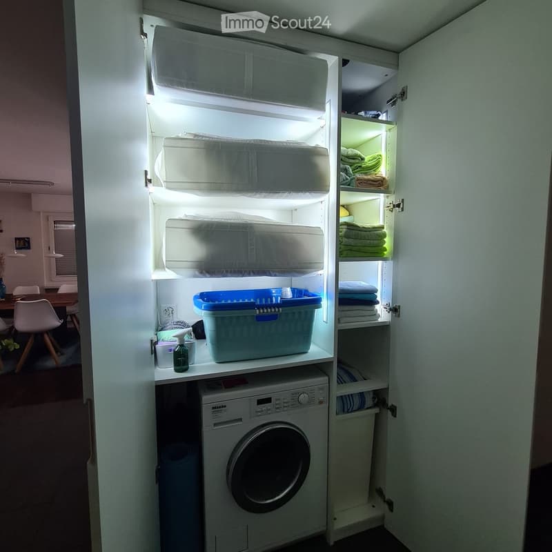 A compact, well-organized laundry area with a built-in washing machine, shelves filled with neatly folded towels, storage bins, and a small seating area with a chair and table visible in the background.