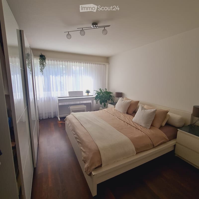 Cozy bedroom with a large bed, nightstand, piano, and decorative plants. The room features wooden flooring and large windows with sheer curtains.