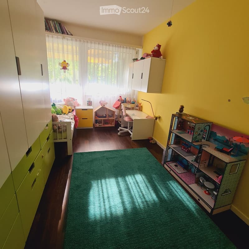 Brightly colored children's bedroom with a single bed, desk, wardrobe, and various toys including a toy house and shelf. Green carpet and large window with white curtains.