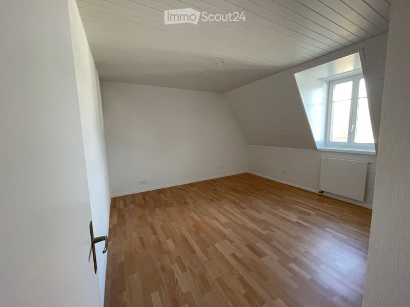 Empty room with wooden flooring, white walls, and a large window with white frame. The ceiling has a slanted design typical of an attic space.