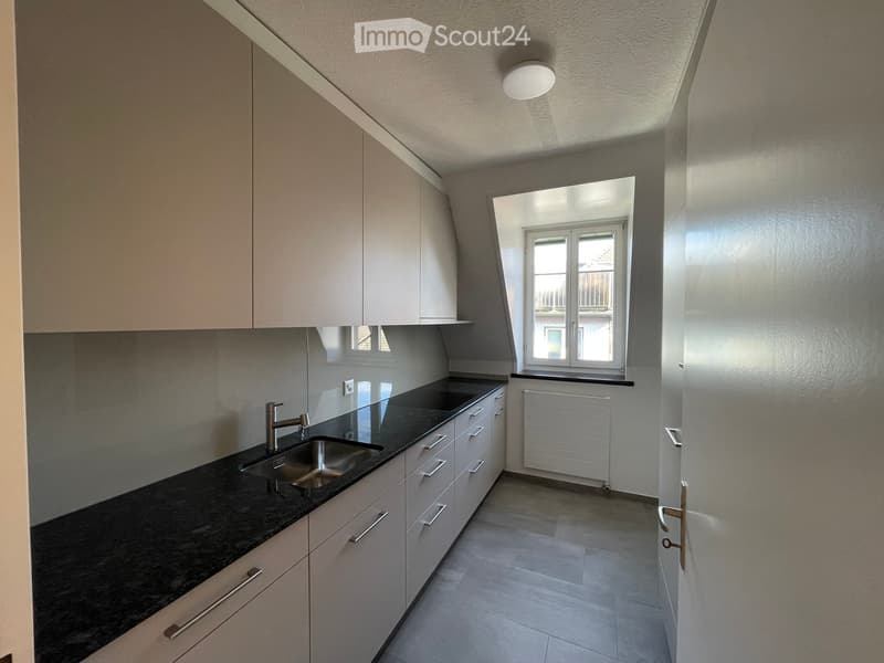 Modern kitchen with sleek black countertops, stainless steel sink, and light-colored cabinets. Features a window with a view and a built-in radiator below.