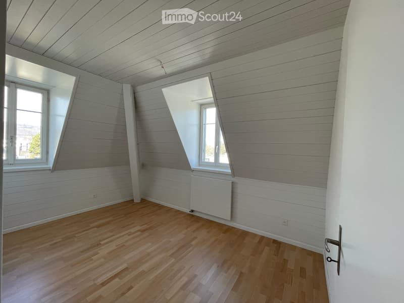 Empty room with wooden flooring, white paneled walls, and slanted ceiling. Two windows with white frames and a door visible.