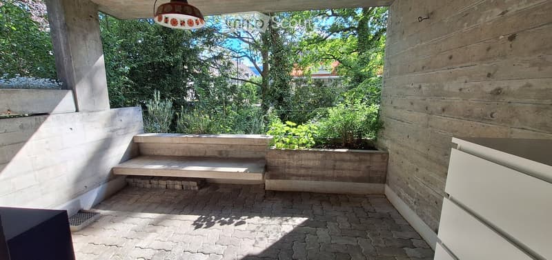 A modern outdoor patio with wooden walls, brick flooring, a wooden bench, and a hanging lantern. The area is surrounded by greenery and has a small raised planter box.