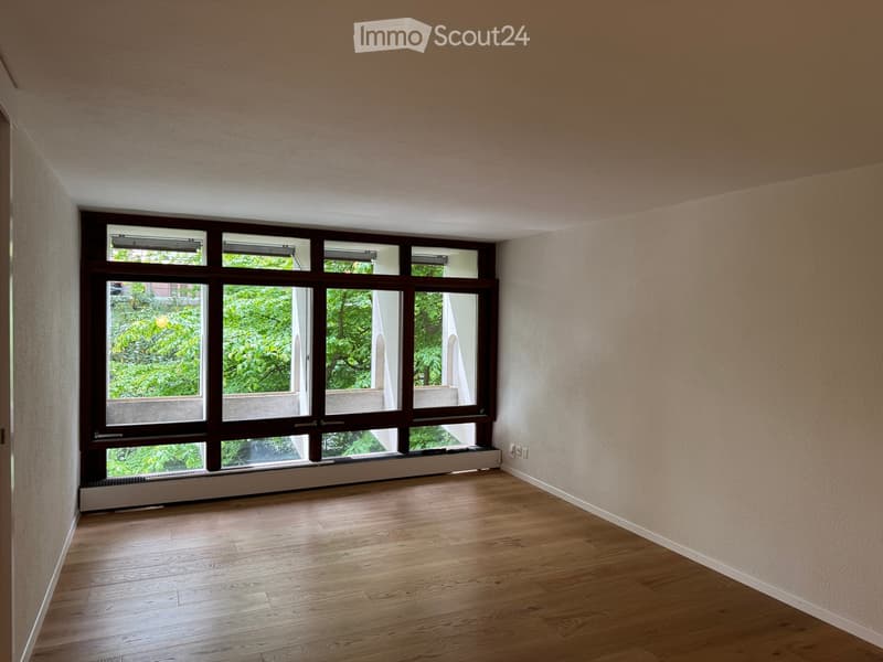 Empty room with wooden flooring and large windows offering a view of greenery outside.