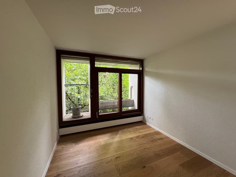 Empty room with wooden flooring, large window with a view of greenery, and a small balcony area visible through the window.