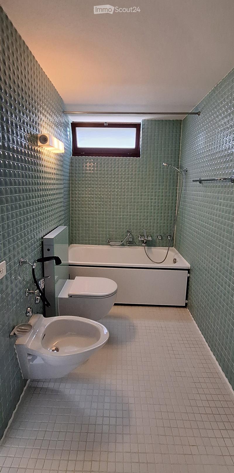 Modern bathroom with green tiled walls, a bathtub, toilet, bidet, and a window above the bathtub.