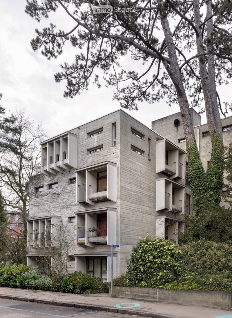 Building. Modernist concrete building with multiple levels, large windows, and balconies, surrounded by trees and greenery.