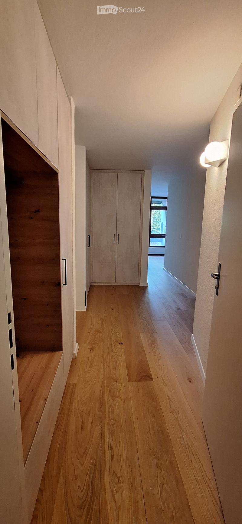 Clean, modern hallway with light wooden flooring, white walls, built-in closet, and a small window at the end.