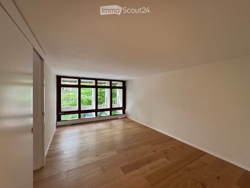 Empty room with wooden flooring and large windows, showing natural light and a view of greenery outside.