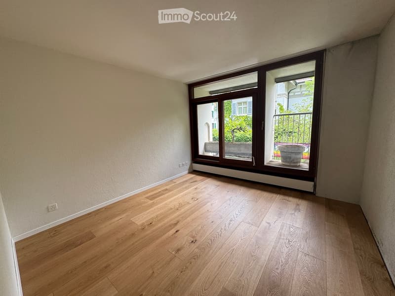 Empty room with wooden flooring and large windows leading to a balcony area. The walls are painted in a light neutral color, and the space appears clean and uncluttered.