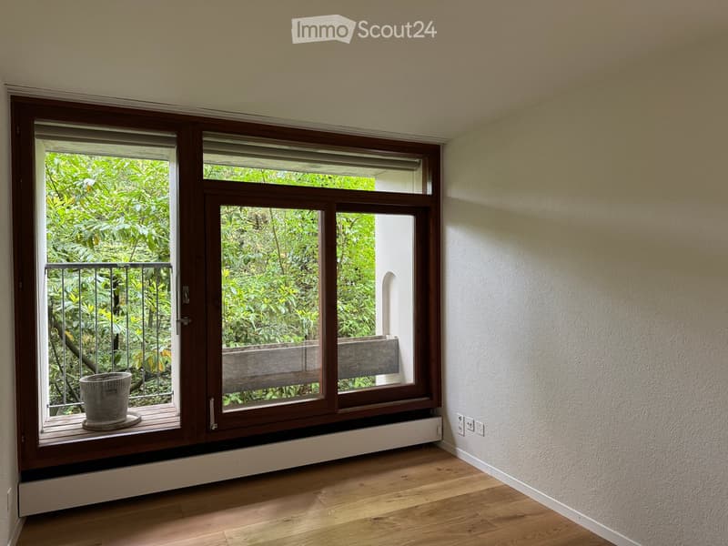 Empty room with wooden flooring, large window with wooden frame and blinds, radiator, and a small balcony area with a bench and potted plant visible through the window.