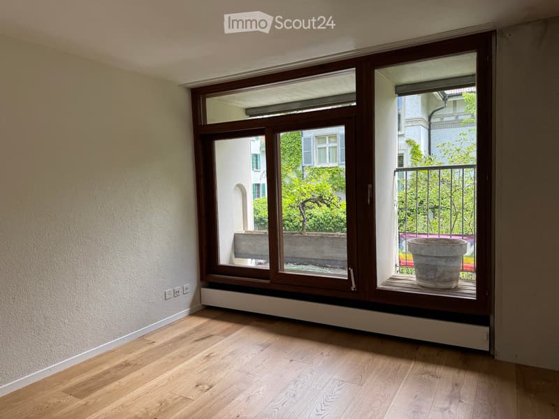 Empty room with wooden flooring, large window with brown frame, radiator under the window, and a view of a balcony with a large pot.