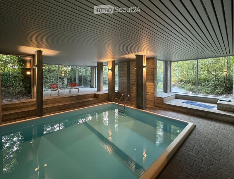 Garden / Outdoor Area. Indoor swimming pool area with large windows overlooking a lush green garden, wooden beams, and a sauna area with a wooden bench and a blue towel.