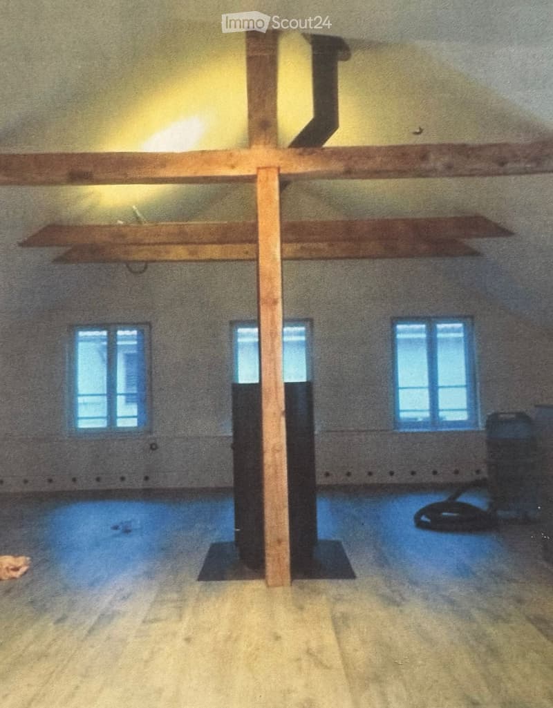 Empty room with exposed wooden beams, concrete walls, and large windows allowing natural light. The floor is concrete, and there are no furnishings or decorations visible.