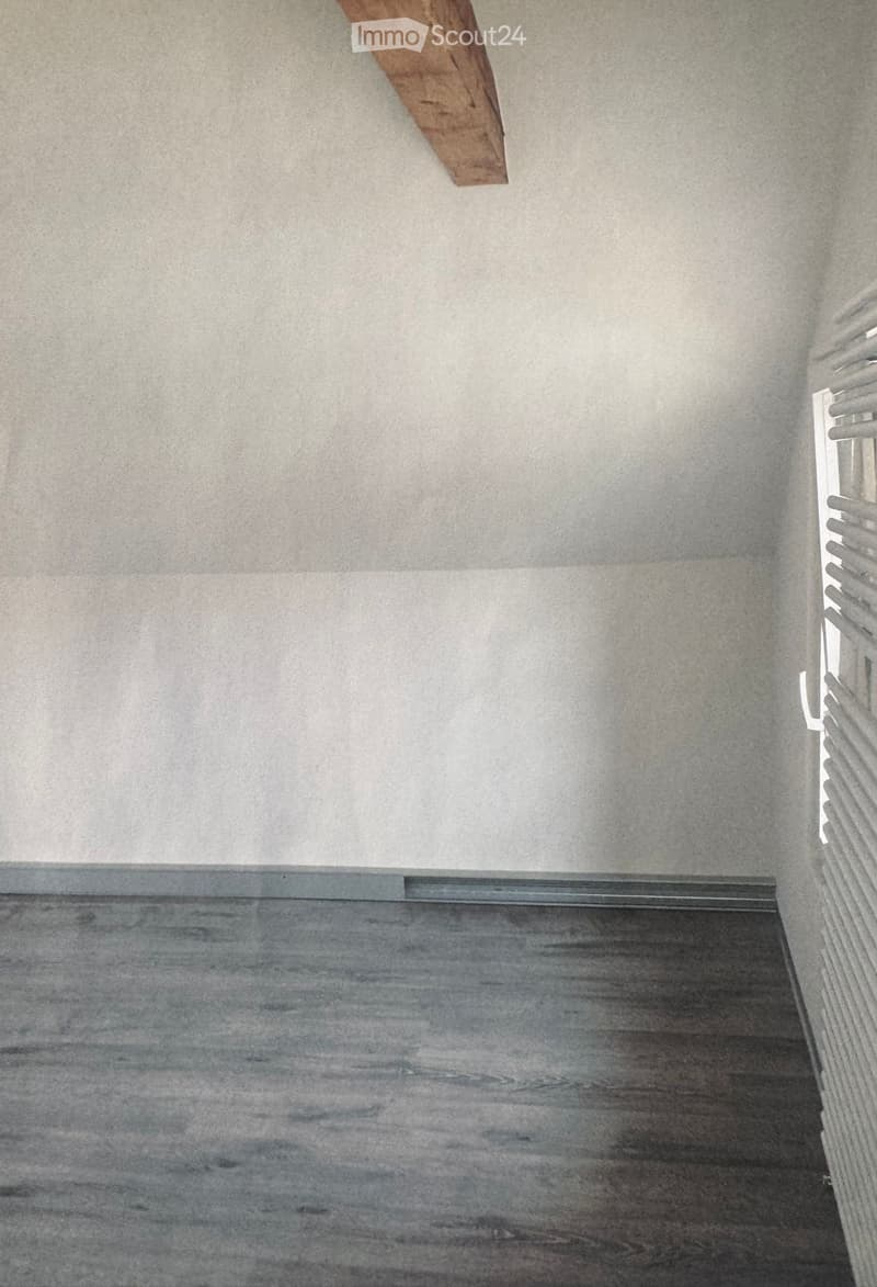 Empty room with grey walls and wooden floor, featuring a small window with white blinds and a wooden beam on the wall.