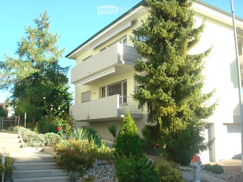 Two-story house with light yellow exterior, multiple balconies, and well-maintained garden with various plants and trees.