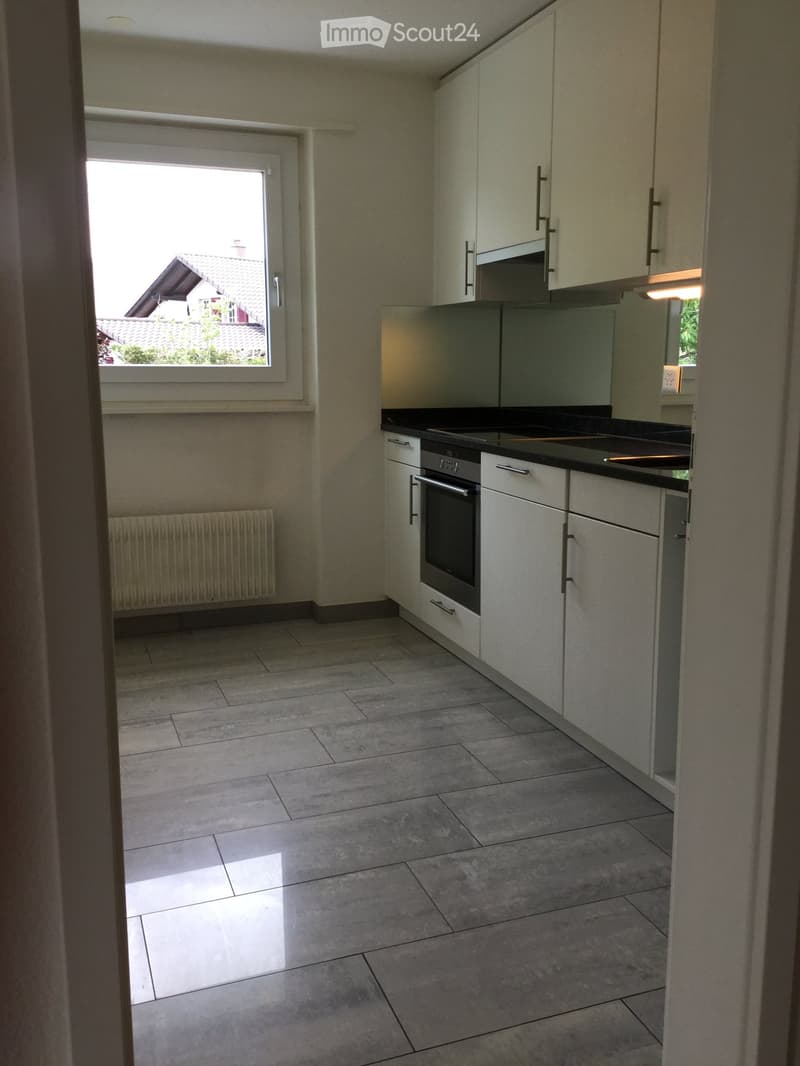 Modern kitchen with white cabinets, black countertop, and tiled floor. Features an oven and a window with a view of a neighboring house.
