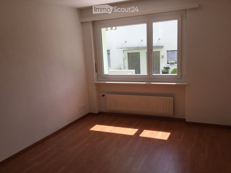 Empty room with wooden flooring, white walls, a radiator, and a large window with a view of a neighboring building and some greenery.