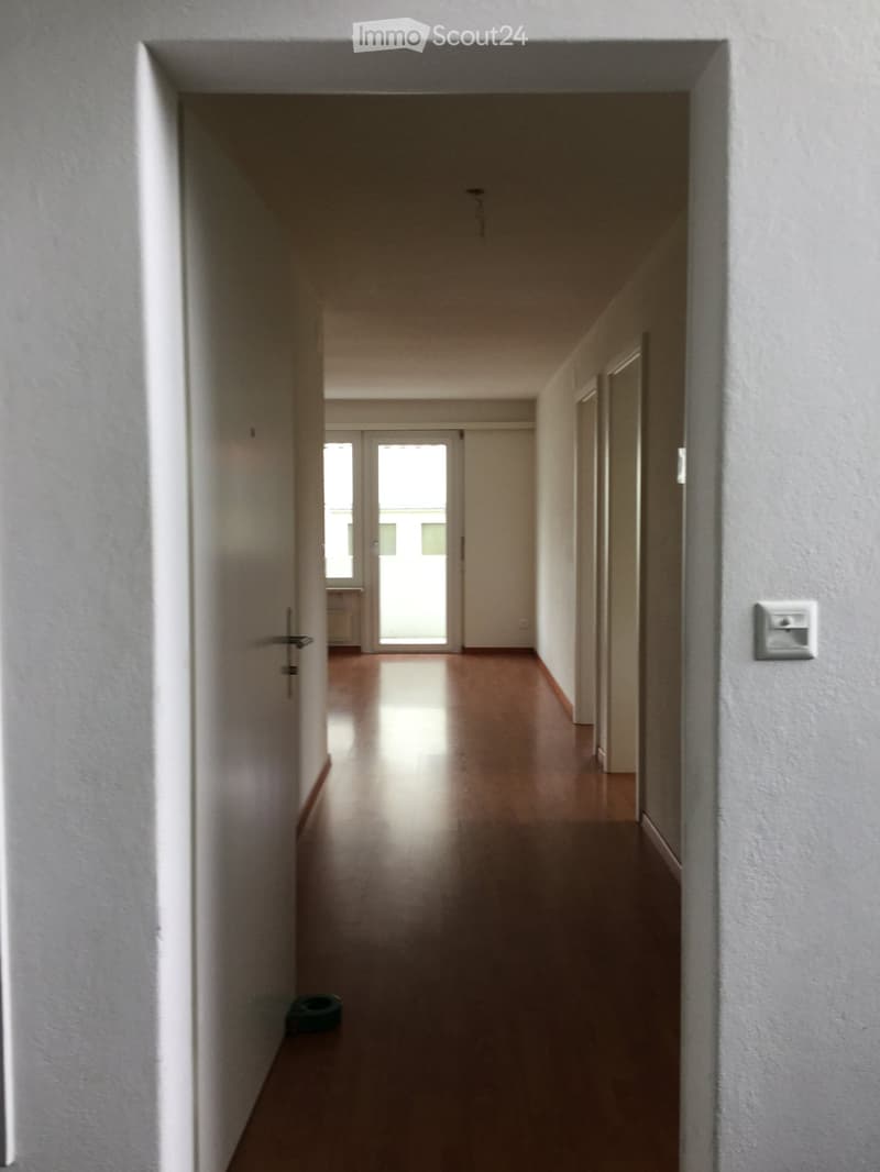 Empty hallway with wooden flooring, white walls, and a door at the end leading to a room with large windows.