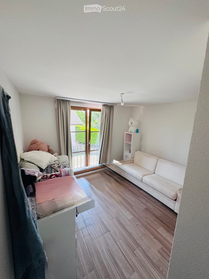 A small, cozy living room with a white sofa, a small table, a shelf with a teddy bear, and a large window with curtains leading to a balcony.
