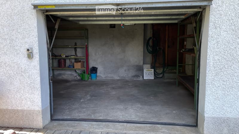 Open garage with concrete floor, metal shelving units, and various stored items including a watering can, buckets, and a coiled hose.