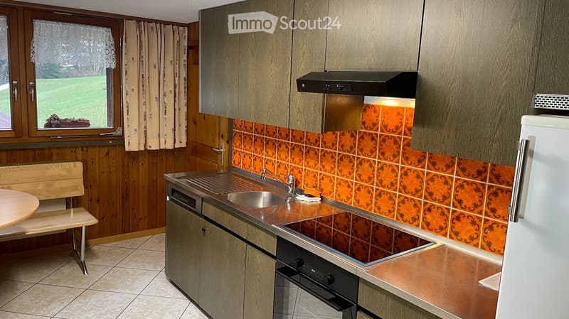 Modern kitchen with wooden cabinets, stainless steel appliances, and orange tiled backsplash. Features a sink, oven, refrigerator, and a small seating area with a bench and table.