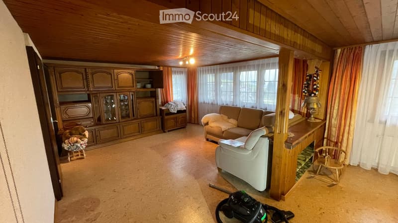 Living room with wooden ceiling, large windows with curtains, sofa, armchair, cabinet, small table, and a vacuum cleaner on the floor.