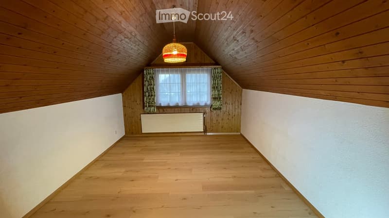 Empty attic room with wooden walls, ceiling, and floor, featuring a single window with curtains and a radiator below it.