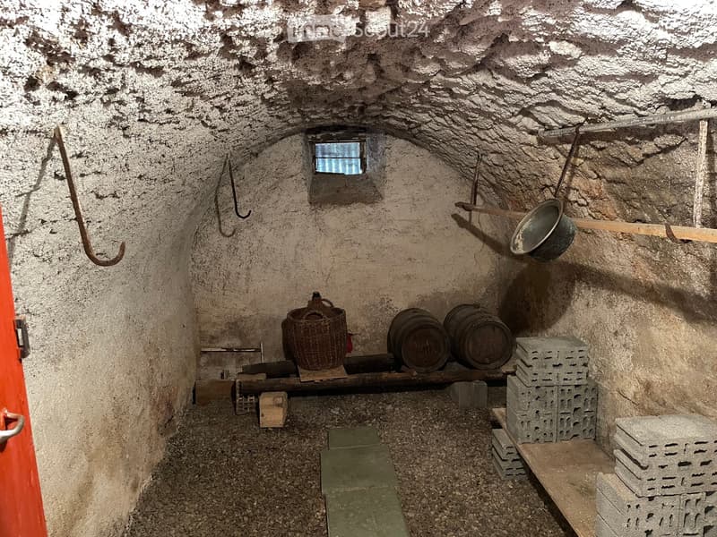 An unfurnished room with rough, textured stone walls and a low, arched ceiling. The space contains a small window, wooden planks, barrels, a metal bucket, and stacks of concrete blocks.