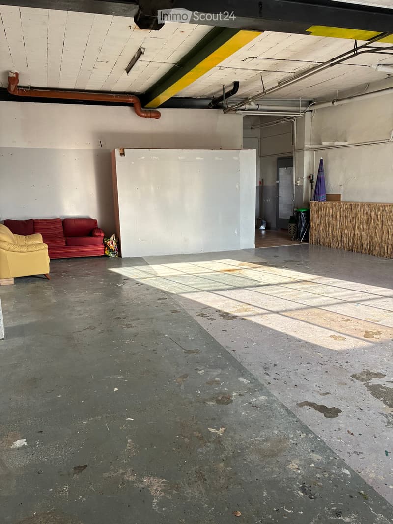 Empty room with concrete floor, white walls, red couch, yellow chair, pipes on ceiling, door, trash bin, wooden counter