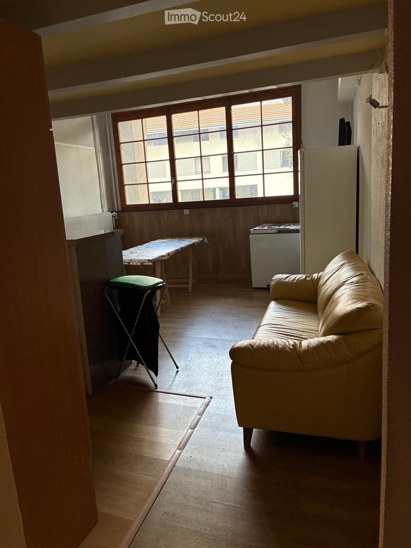 A room with a couch, a table, a stool, a refrigerator, and a window