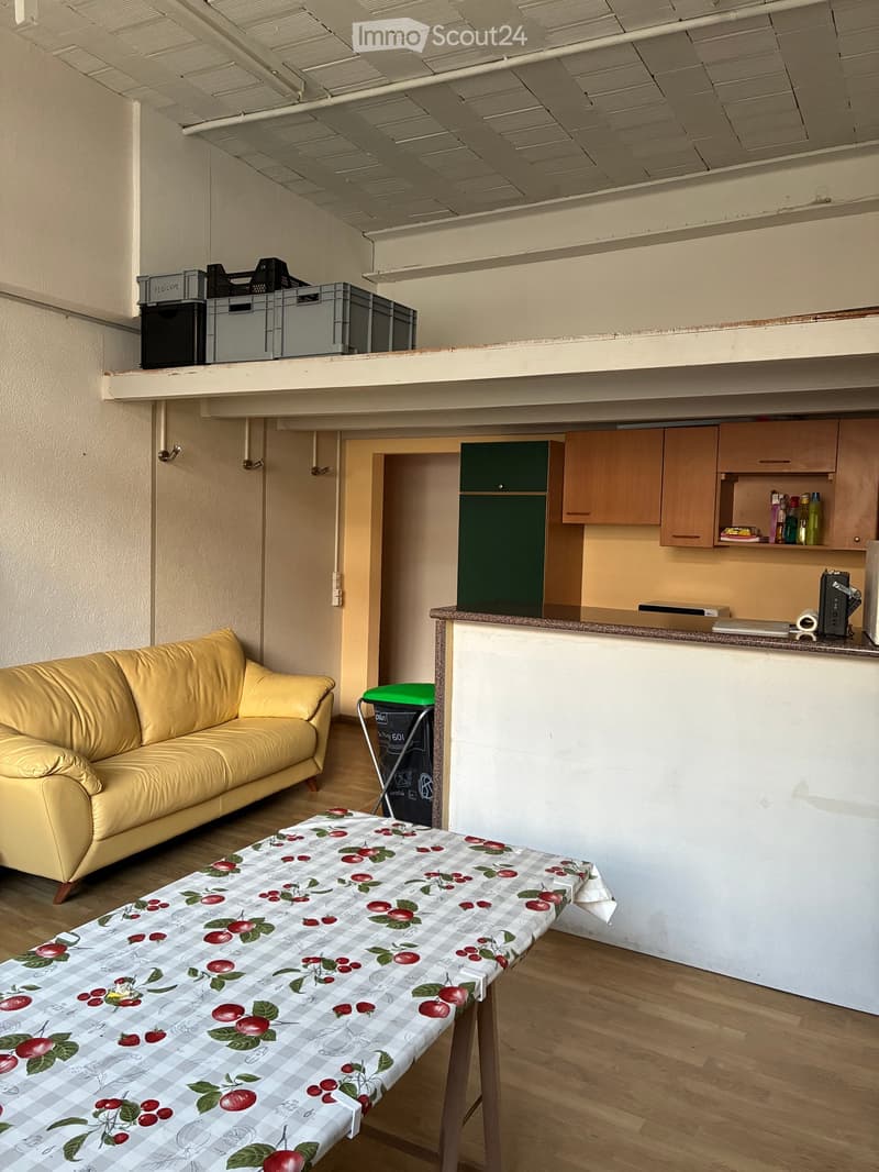 A room with a yellow couch, a table with a floral tablecloth, a kitchen with wooden cabinets, a countertop, and a trash bin, and a storage box on the wall.