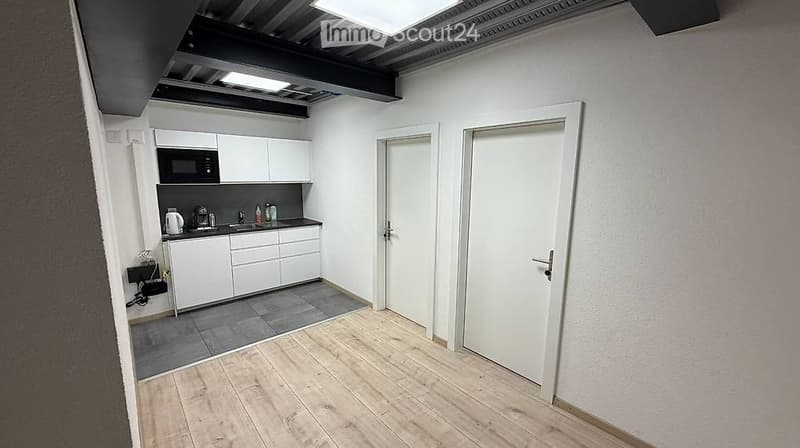 The image shows a modern, minimalist kitchen area connected to an open living space. The kitchen features white cabinets, a microwave, a sink with a faucet, and a coffee maker on the counter. The living area has light wooden flooring and white walls, with