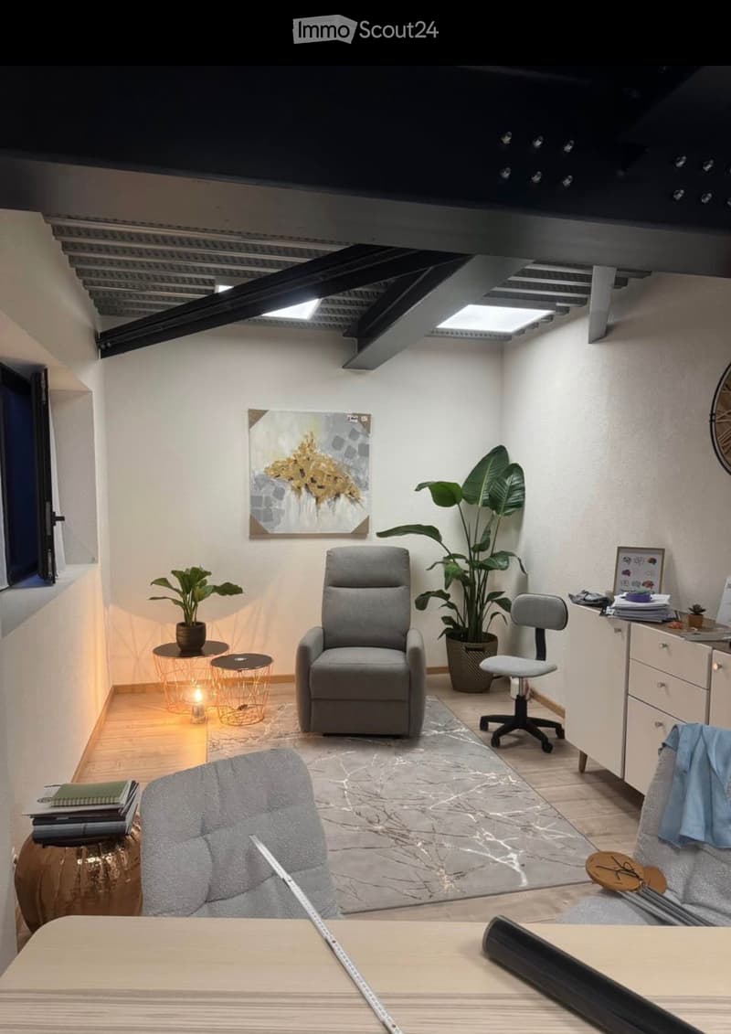A modern, well-lit room with a mix of furniture including a grey armchair, a desk, a chair, a small round table, and a potted plant. The room features a large painting on the wall and a sleek, contemporary design with wooden flooring and white walls.