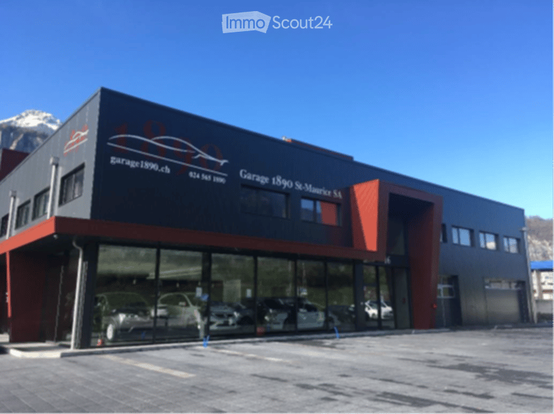 Modern commercial building with large glass windows and a red and black facade, featuring signage for "Garage 1890 St-Maurice SA" and a spacious parking area.