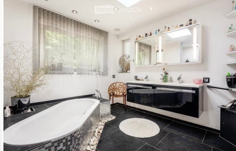 Modern bathroom with a freestanding bathtub, double sink vanity, large mirror, wicker chair, and decorative plant. Features black tiled flooring and white walls.