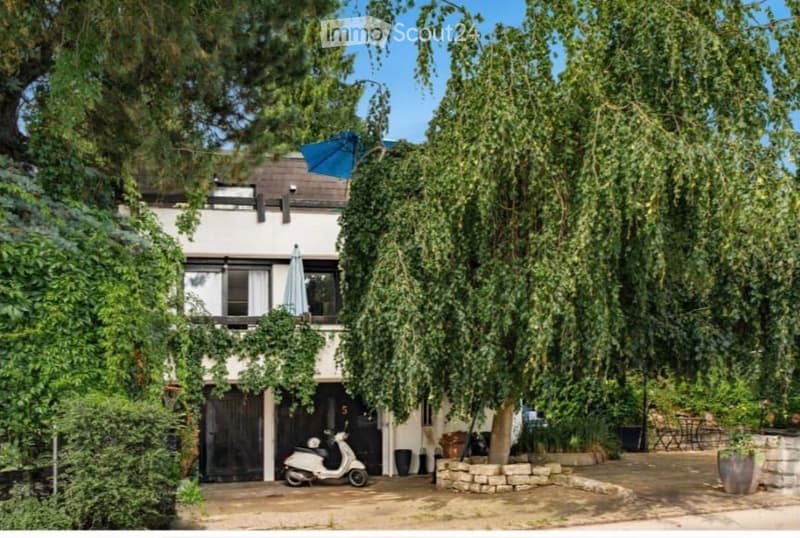 Two-story modern house with white exterior, large windows, and a covered garage entrance. A scooter is parked in the garage, and the house is surrounded by lush greenery and trees.