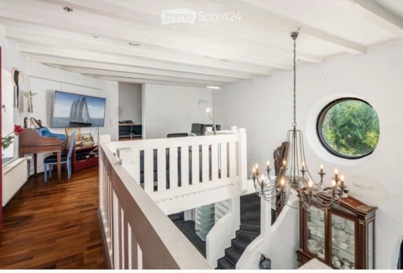 Spacious, well-lit hallway with wooden flooring, white walls, and a staircase leading upwards. Features a chandelier, a round window, and a piano in the adjacent room.