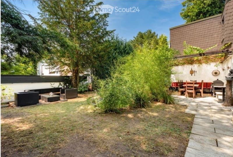 A well-maintained garden with a mix of greenery, including trees and shrubs, a paved pathway, outdoor seating area with a table and chairs, and a small lawn area.