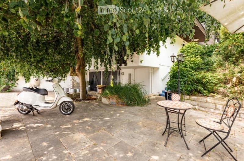 A shaded outdoor patio area with a stone floor, a small round table with two chairs, a white scooter, and a small dog sitting on the scooter. The area is surrounded by lush greenery and a white building with large windows.