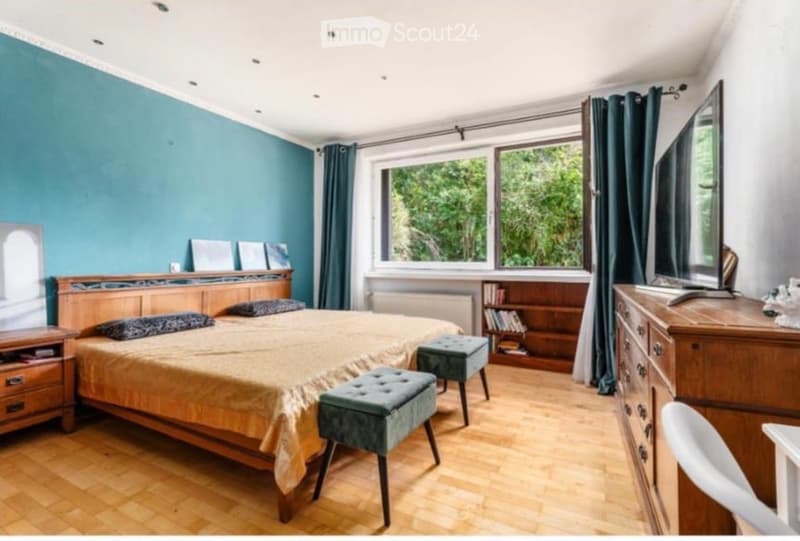 Cozy bedroom with wooden flooring, teal accent wall, large window with green curtains, bed with patterned bedding, nightstand, wooden bookcase, and a mirror on the dresser.