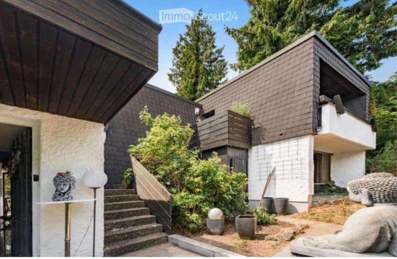 Modern two-story house with a raised ground floor, featuring a balcony, wooden roof, and surrounding greenery.