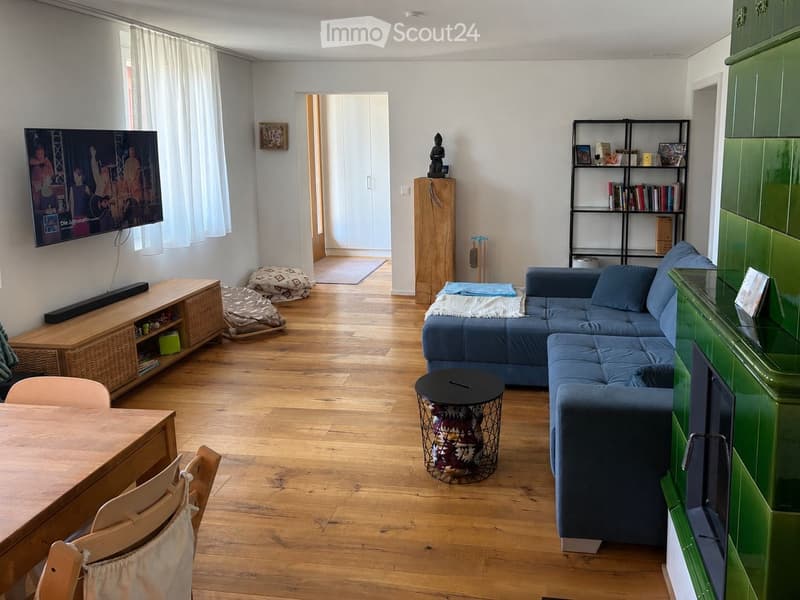 A modern living room with wooden flooring, a blue sectional sofa, a wooden dining table with chairs, a TV on a wooden stand, a bookshelf, and decorative items including a small statue and a woven basket.
