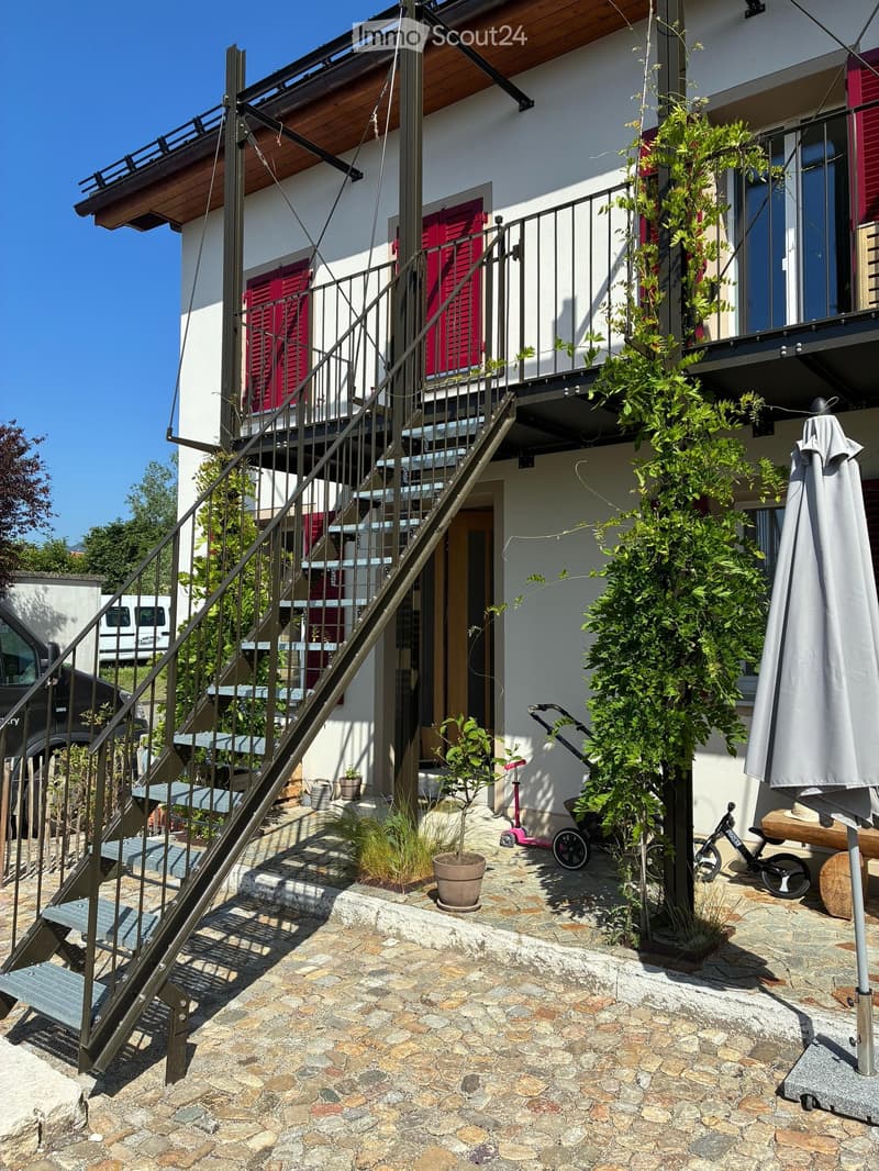 Outdoor area with stone-paved ground, staircase leading to a balcony, potted plants, and a closed umbrella.