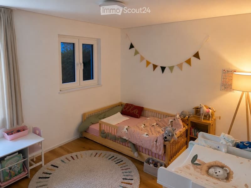 A cozy children's bedroom with a wooden toddler bed, a crib, a nightstand with toys, a small table with storage, and a decorative rug on the floor.