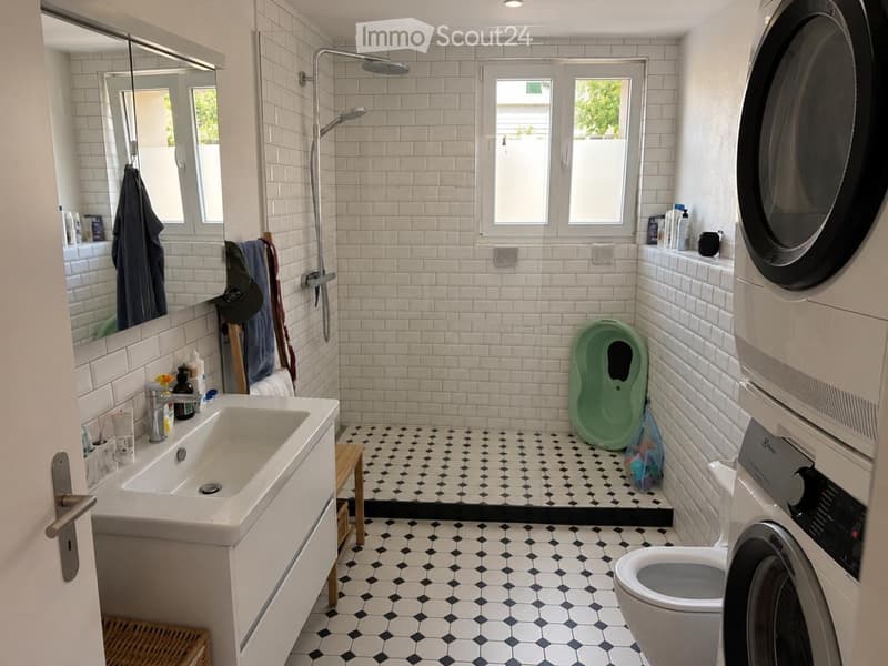 Modern bathroom with white tiled walls, black and white checkered floor, sink, toilet, washing machine, shower, and a small green bath tub.