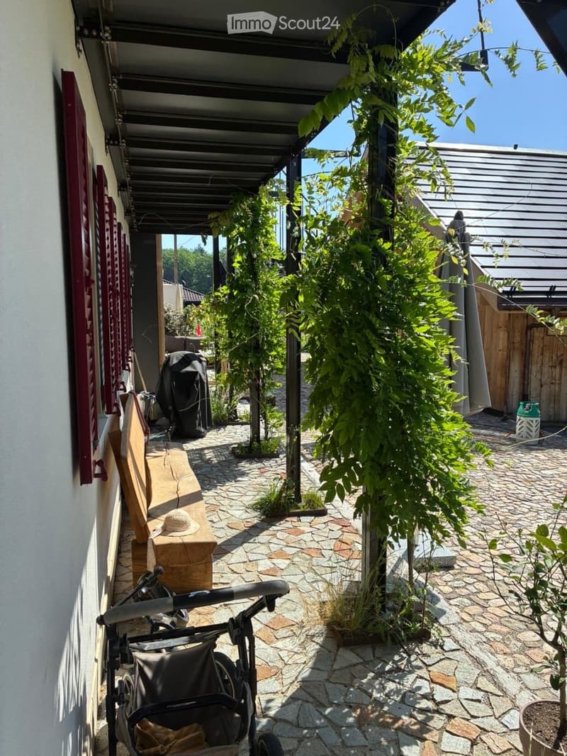 A covered outdoor area with a stone-paved walkway, greenery, and a stroller parked near the entrance.
