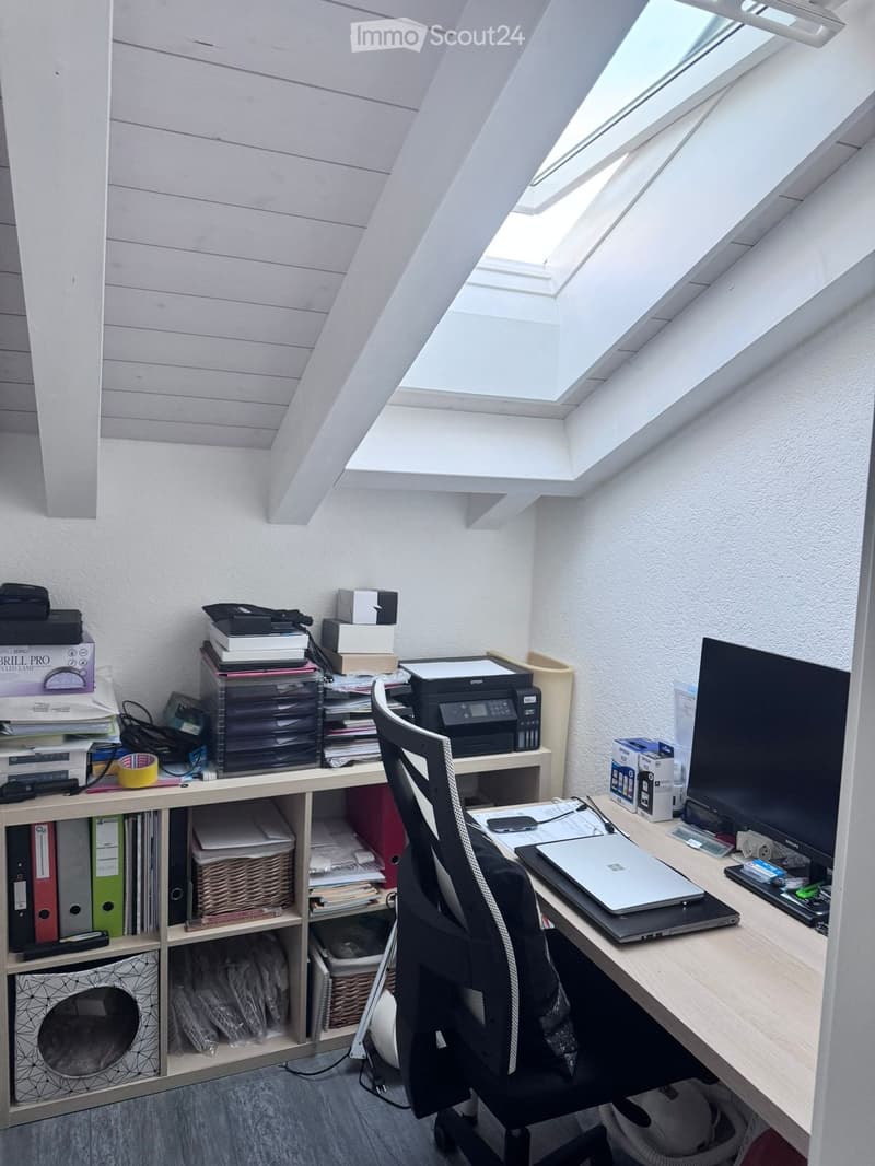 A well-lit attic office with a large skylight, featuring a wooden desk with a laptop, a black office chair, a printer, and shelves filled with books, folders, and office supplies.