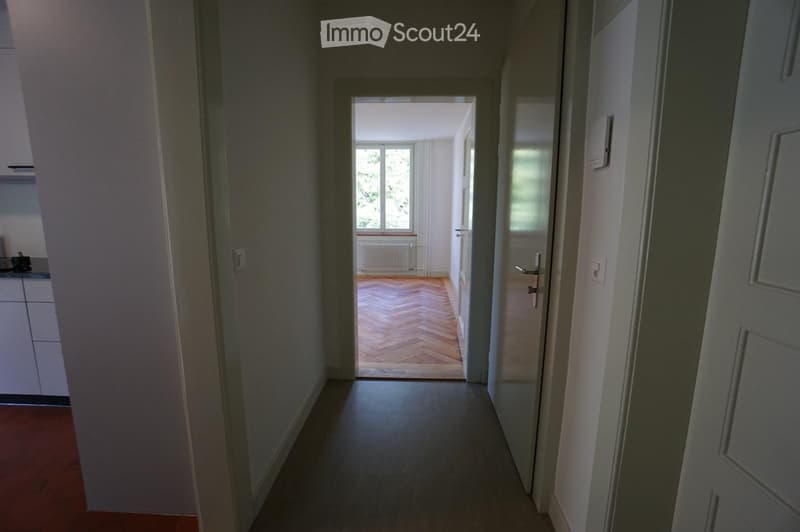 A narrow hallway with a polished floor, white walls, and a door leading to a room with wooden flooring and a window. The hallway has a radiator and a small cabinet visible on the left side.