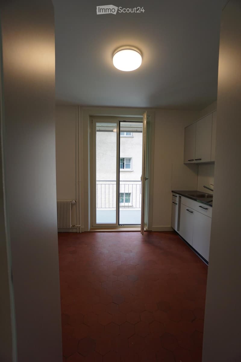 Empty hallway with red hexagonal tiles, white walls, a round ceiling light, and an open door leading to a balcony area.